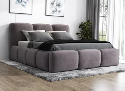 A modern upholstered bed with a grey fabric finish, set up in a bedroom with artwork on the wall and curtains in the background.