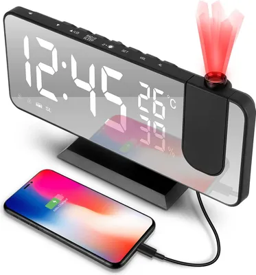 A digital alarm clock with a projected display and a smartphone charging on its base.