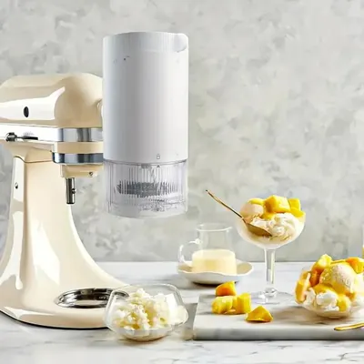 A cream-colored stand mixer with a white ice shaver attachment, accompanied by bowls of ice cream and chopped mangoes.