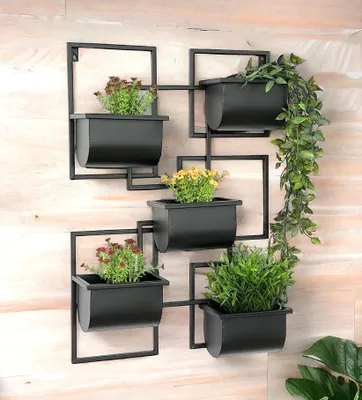 A black metal wall-mounted planter with multiple pots holding plants, displayed against a light wood-paneled wall.