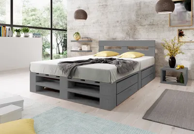 A bedroom with a grey wooden pallet bed frame with storage drawers, a mattress, pillows, and a blanket. There are shelves on the wall, a nightstand with a vase of flowers, and a large window with a view of trees.