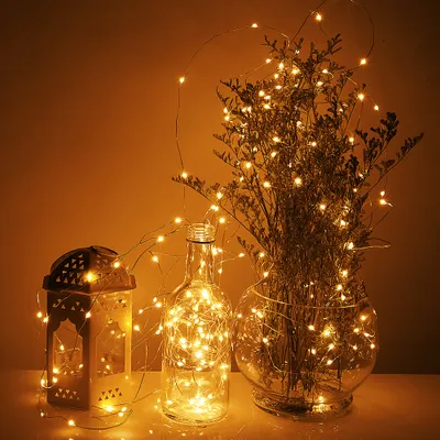 A decorative arrangement of fairy lights in a bottle and a lantern, with dried flowers in a vase.