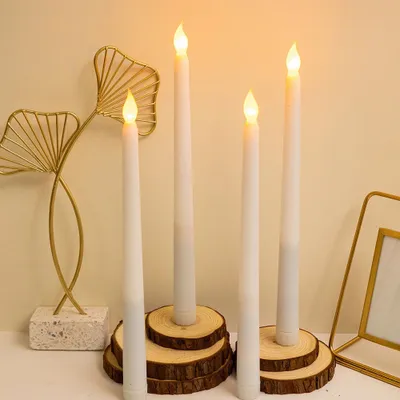 Four LED taper candles are displayed on a surface, with decorative golden leaf-like objects and a picture frame in the background.