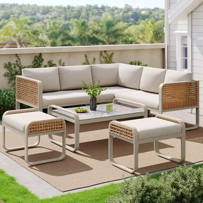 An outdoor patio set with a sectional sofa, coffee table, and ottomans on a rug, with greenery and a building in the background.