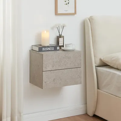 A floating nightstand with two drawers is mounted on a white wall next to a bed. The nightstand has a concrete-like texture and is decorated with a candle, a book, a vase with flowers, and a cup of coffee.