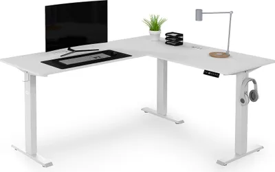 A white L-shaped standing desk with a monitor, keyboard, mouse, desk lamp, and headphones.