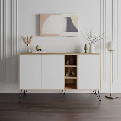 A modern sideboard with white doors and a natural oak-effect frame, standing on black hairpin legs. It's styled with decorative items and a piece of abstract art hangs on the wall above it. A floor lamp is positioned to the right.