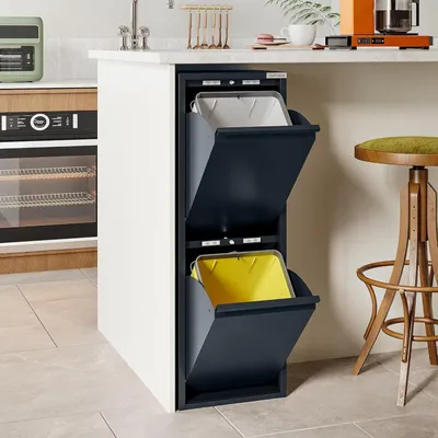 A modern kitchen scene with a built-in trash can unit featuring two compartments, one with a white liner and the other with a yellow liner. A stool and kitchen appliances are visible in the background.