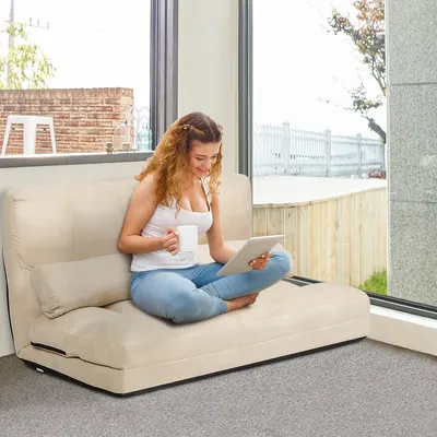 A woman is sitting on a beige sofa bed, holding a tablet and a mug, in a well-lit room with a large window overlooking an outdoor area.
