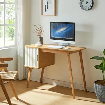 A desk with a computer and a plant in a room with a window.