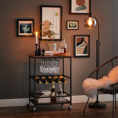 A bar cart is set up in a room with artwork on the wall and a chair nearby.