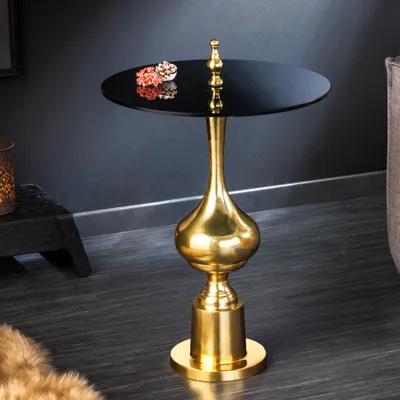A gold and black accent table with decorative items on top, placed on a dark wooden floor next to a dark wall and a furry rug.