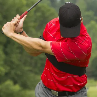 A golfer is shown mid-swing, wearing a red shirt and a black cap. A black arm brace is visible around their torso.