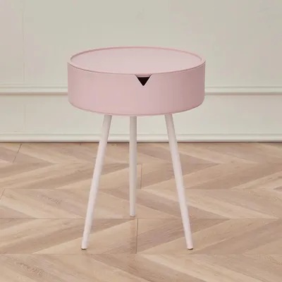 A pink round side table with three white legs is shown against a white and light gray wall.