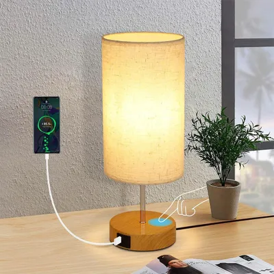 A bedside lamp with a phone charging on it, placed on a wooden desk next to a plant.