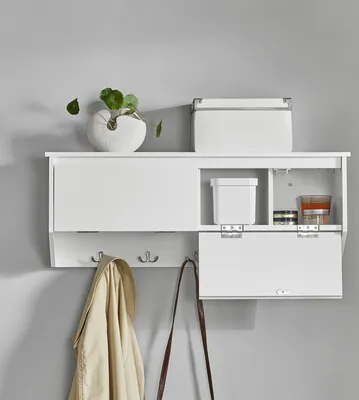 A white wall-mounted storage unit with hooks below, holding a jacket and a bag. Above, on the shelf, are a decorative vase with a plant and a storage box. One of the lower compartments of the unit is open, revealing interior storage and a small container.