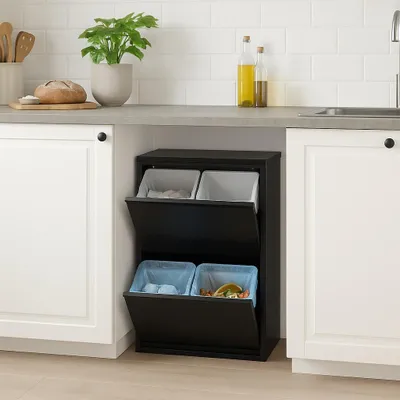 A black multi-compartment trash can with two flip-down doors, situated under a kitchen counter between two white cabinets. The trash can contains four bins, two at the top and two at the bottom, with blue liners in the bottom bins.
