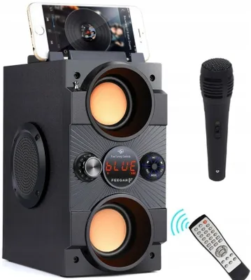 A black portable Bluetooth speaker with a smartphone docked on top, a microphone to the right, and a remote control at the bottom right.