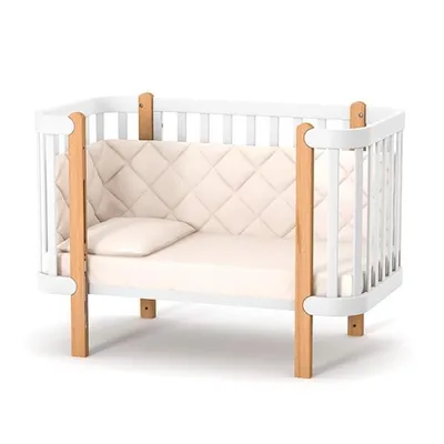 A white baby crib with wooden accents and a light-colored mattress and bumper pad, shown against a white background.