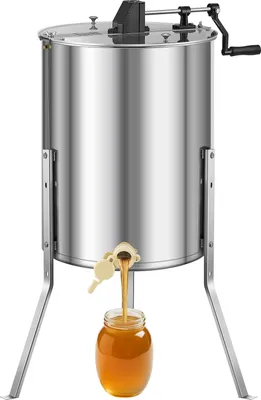 A stainless steel honey extractor with a crank handle and a spigot, dispensing honey into a glass jar.