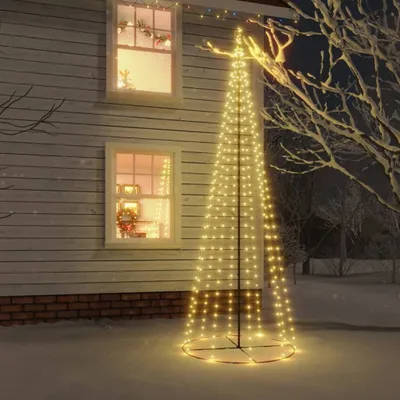 A conical LED Christmas tree decorated with warm white lights stands in the snow next to a house with illuminated windows.