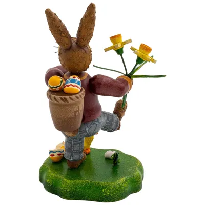 Ein fröhlicher Hase in bunter Kleidung trägt einen Korb voller Ostereier auf dem Rücken und hält einen Strauß gelber Narzissen in der Hand während er auf einer grünen Wiese steht