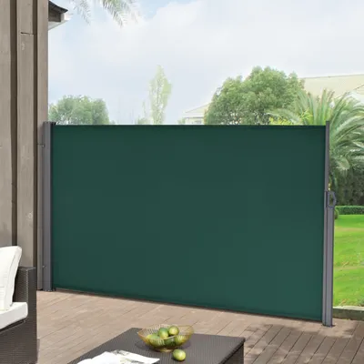A dark green retractable side awning is extended on a wooden deck, providing privacy and shade. The awning is attached to a wooden post on the left and a gray metal pole on the right. In the background, there is a green lawn and trees under a cloudy sky. A