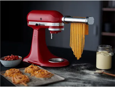 A red KitchenAid stand mixer with a pasta attachment is making fresh pasta. There is a bowl of chopped tomatoes and a jar of flour in the background, with freshly made pasta on a tray in the foreground.