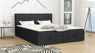 A dark gray boxspring bed with white bedding and pillows is shown in a bedroom setting with a patterned rug and a floor lamp.