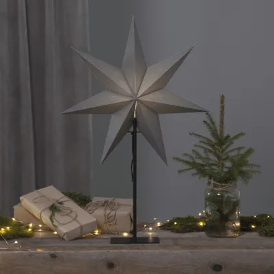 A large, grey, star-shaped lamp is displayed on a wooden surface, surrounded by wrapped gifts and small fairy lights. A miniature Christmas tree in a glass jar is also present, creating a festive, indoor holiday scene.