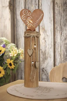A decorative wooden column with a metal heart topper, adorned with twine and wooden hearts, placed on a table with flowers in the background.