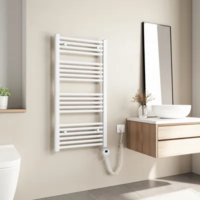 A white electric towel radiator is mounted on a light beige wall in a modern bathroom, next to a vanity with a sink and mirror.