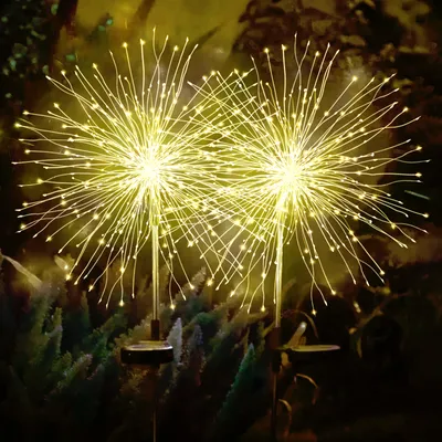 Two solar-powered LED garden lights resembling fireworks are illuminated at night, set amongst dark green foliage.
