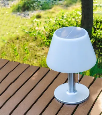 A solar table lamp sits on a wooden deck outdoors, with green grass and a tree in the background.