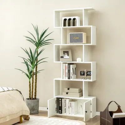 A white, modern, zig-zag bookshelf filled with decorative items and books, placed next to a large potted plant and a bed in a room.