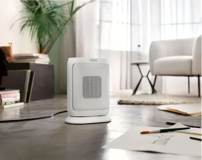A white De'Longhi ceramic heater is placed on a grey floor in a living room setting, with a chair and a plant in the background.