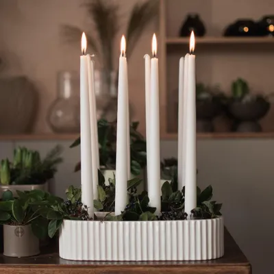 A white ribbed candle holder with five white candles and greenery on a wooden table.