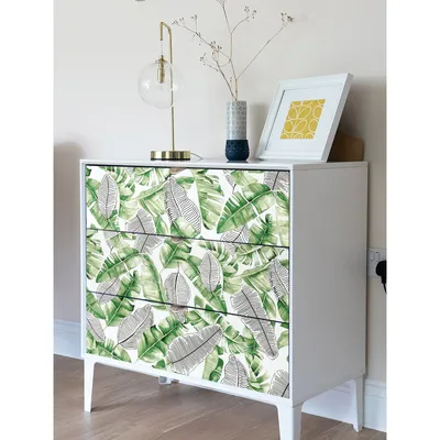 A white dresser with drawers decorated with a tropical leaf pattern, a gold lamp, and a framed picture on top.