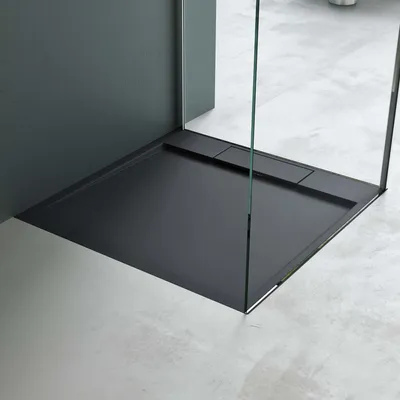 A modern, minimalist shower tray with a dark gray finish, set against a light gray floor and a dark gray wall. A clear glass shower screen is visible.