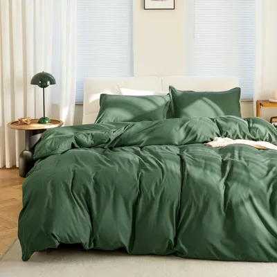 A neatly made bed with dark green bedding in a room with white curtains and blinds.