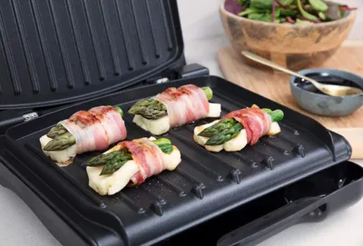 Asparagus and cheese wrapped in bacon are being grilled on a George Foreman grill. A salad and a bowl of sauce are visible in the background.