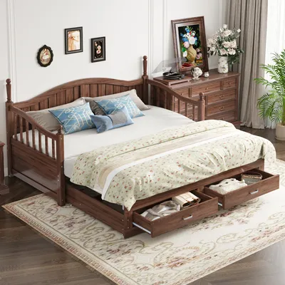 A wooden daybed with drawers underneath, dressed with bedding and pillows, in a room setting with a dresser and plant.