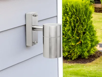A silver outdoor wall light mounted on a light gray, horizontally sided wall, with a green bush and grass in the background.