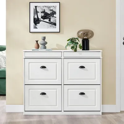 A white shoe cabinet with four flip-down drawers is shown against a cream-colored wall. Decorative items, including two vases and a lamp, are placed on top of the cabinet.