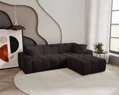 A dark gray sectional sofa is placed on a light-colored rug in a modern living room. The wall behind the sofa features a textured, wavy design in shades of gray and beige. A large abstract artwork is on the left, and a small side table with a plant is on t