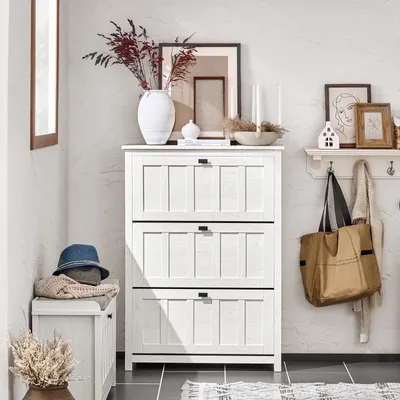 A white shoe cabinet with three flip-down drawers, decorated with framed pictures, a vase with dried flowers, and a tote bag hanging on a shelf.