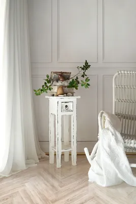 A white side table with a drawer and decorative items on top, placed next to a white chair with a blanket draped over it, in a room with white paneled walls and sheer white curtains.