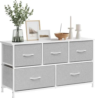 A white dresser with five gray fabric drawers and white handles, decorated with a vase of greenery, two framed pictures, and two candle holders with candles.