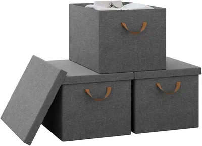 Three grey fabric storage boxes with brown handles are arranged on a white background. One box is stacked on top of two others, and its lid is propped open, revealing a white shirt inside. The lid of one of the bottom boxes is also open and leaning against
