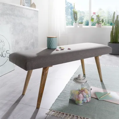 A grey upholstered bench with wooden legs is placed on a rug in a room with a window in the background.